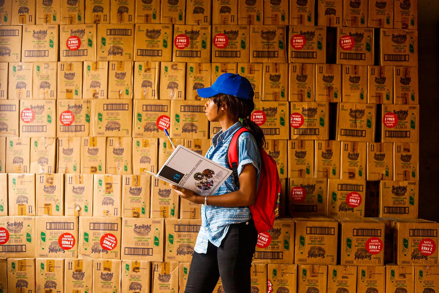A worker with a clipboard checking stock against a wall of stacked cartons