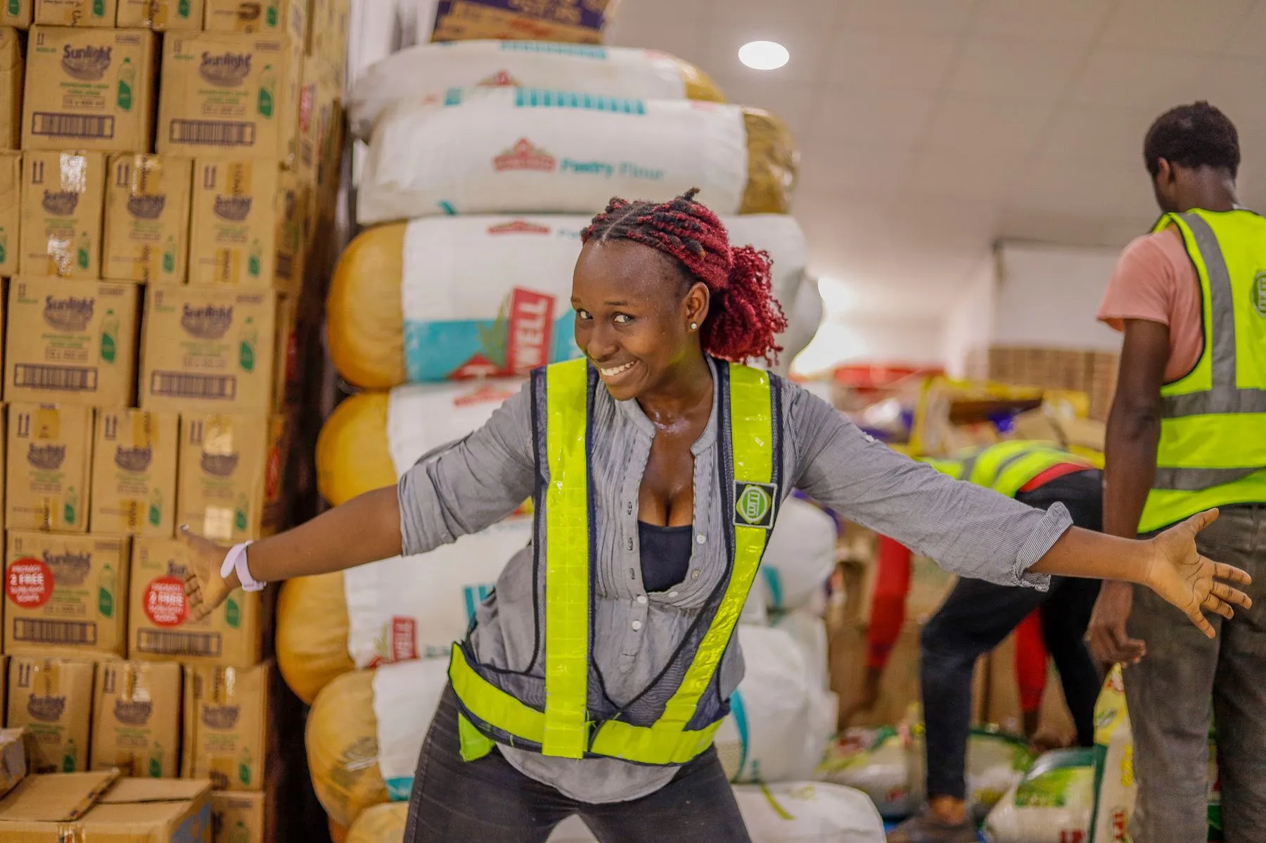 A warehouse worker in a hi-vis vest smiling beside stacked cartons and sacks of flour