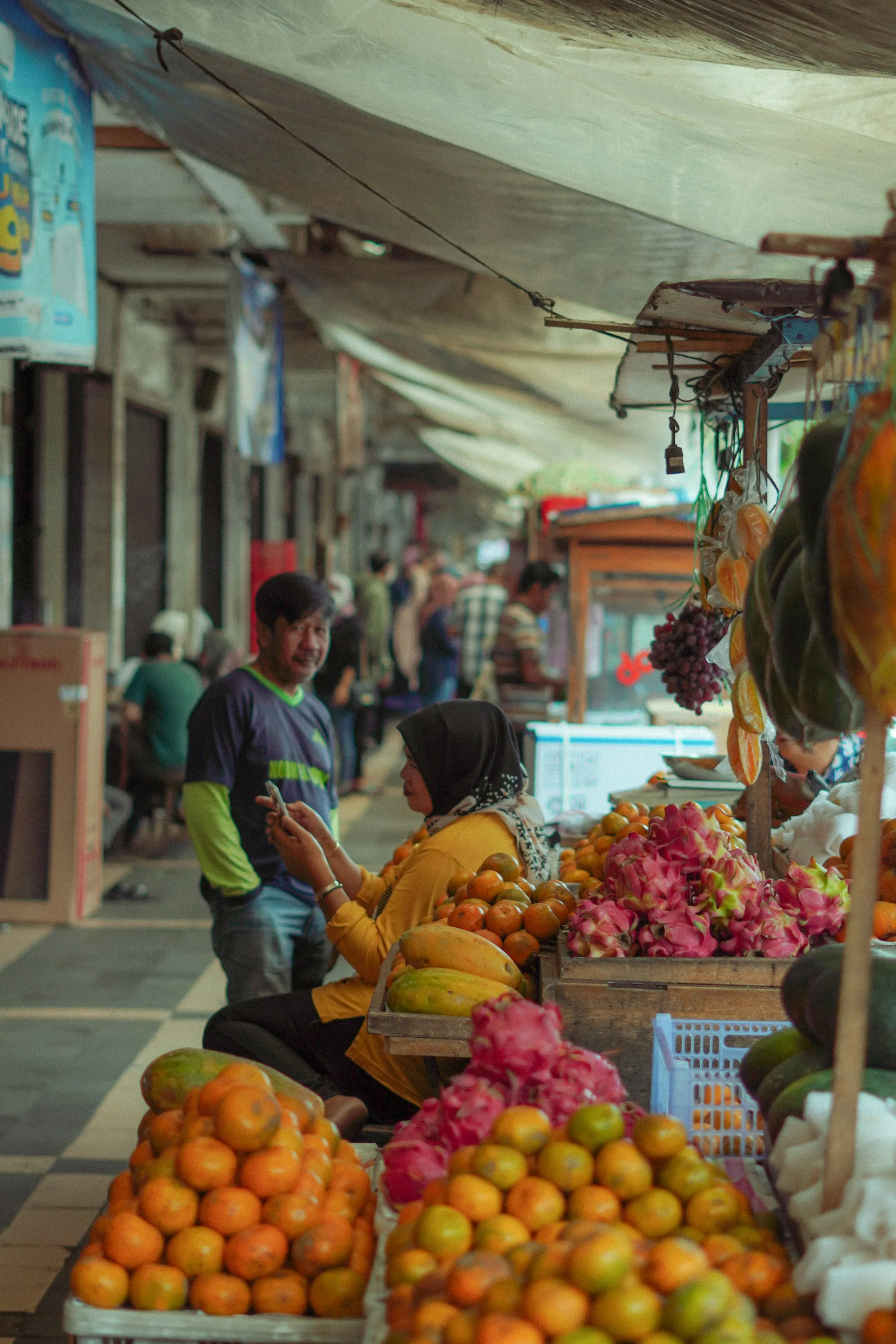 A trader at a busy market stall checks his phone while talking with a customer beside crates of fresh fruit