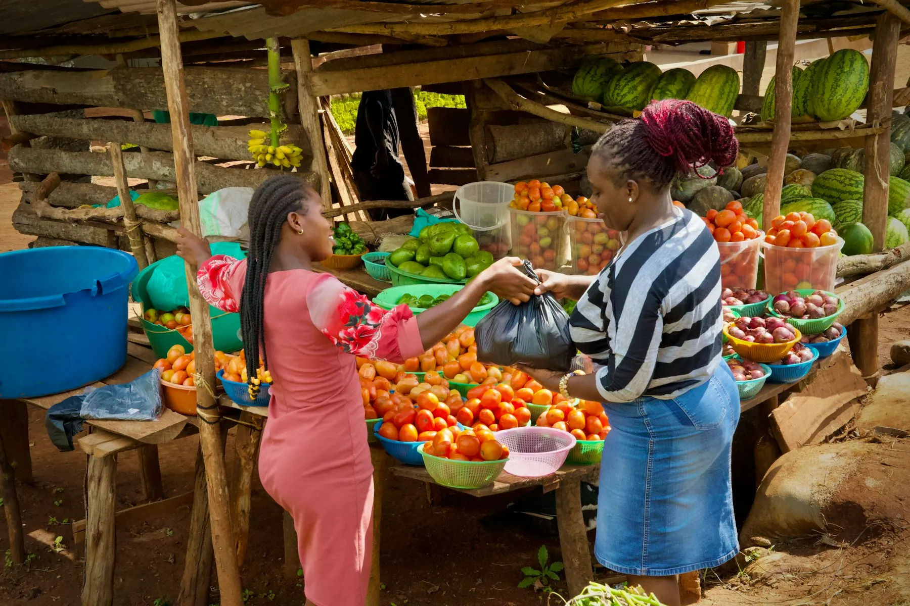 A vendor handing a customer a bag of produce at an open-air fruit and vegetable stall