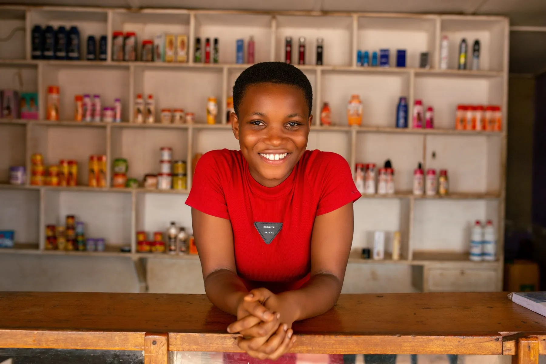 A smiling young shop owner leaning on her counter in front of stocked shelves